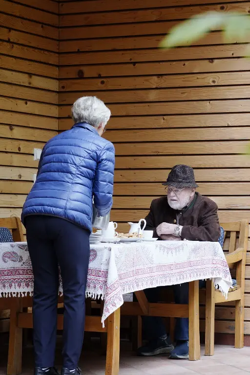 Älterer Herr sitzt am Tisch auf der Terasse und eine ältere Dame steht nebenbei und richtet Getränke