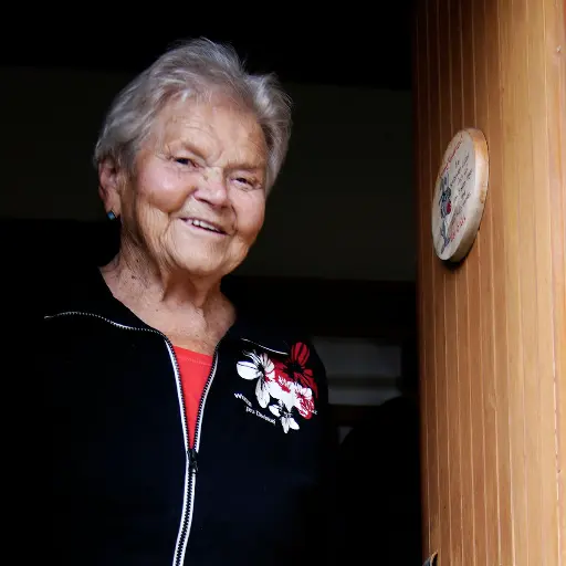 Pensionisting steht bei sich in der Wohnung und lächelt in die Kamera.