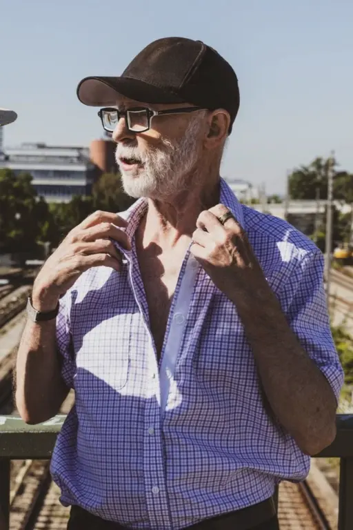 Älterer Mann mit grauem Bart, schwarzer Kappe und Brille steht im Freien, hat die Hände am Hemdkragen und blickt zur Seite.